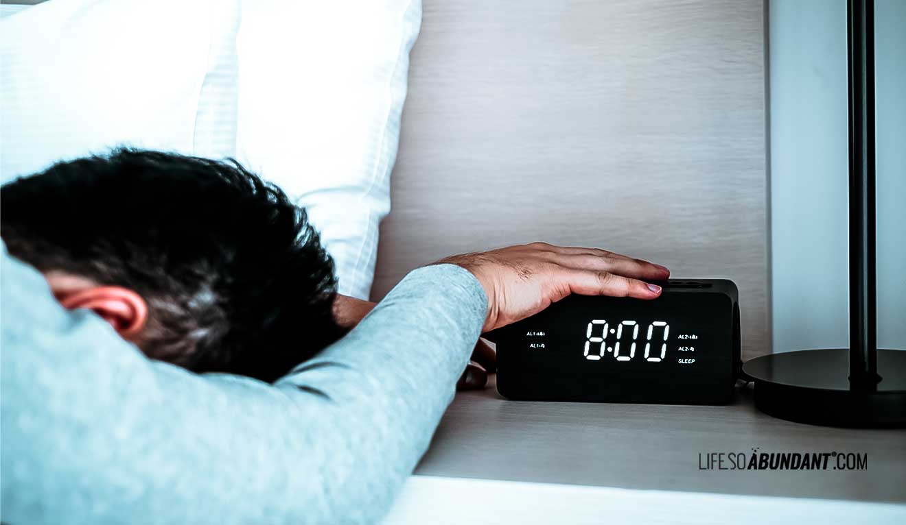 Man reluctant to come out of bed hand on an alarm clock on a nightstand - Life So Abundant®