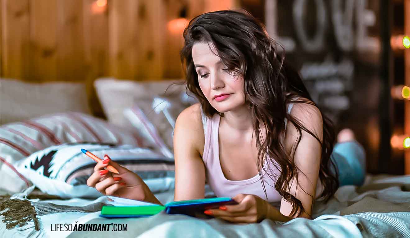 Pretty woman in bed pen in hand looking over notes in her notebook - Life So Abundant®
